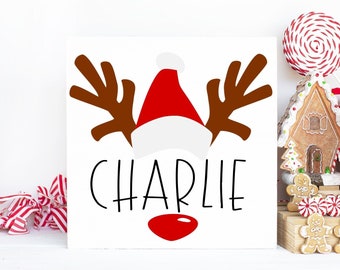 May include: A white wooden sign with a red and white Santa hat, brown reindeer antlers, and the name "CHARLIE" in black script.