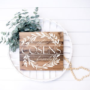 May include: A rustic wooden sign with the text "The Cosens family" in white script, framed by a white floral wreath. The sign is set against a white background with eucalyptus and a beaded garland.
