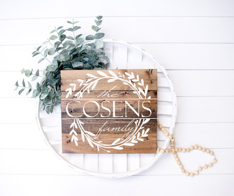 May include: A rustic wooden sign with the text "The Cosens family" in white script, framed by a white floral wreath. The sign is set against a white background with eucalyptus and a beaded garland.