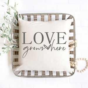 May include: A white throw pillow with the text "LOVE grows here" in gray lettering with a small heart symbol. The pillow is in a woven basket with a wooden bead garland.