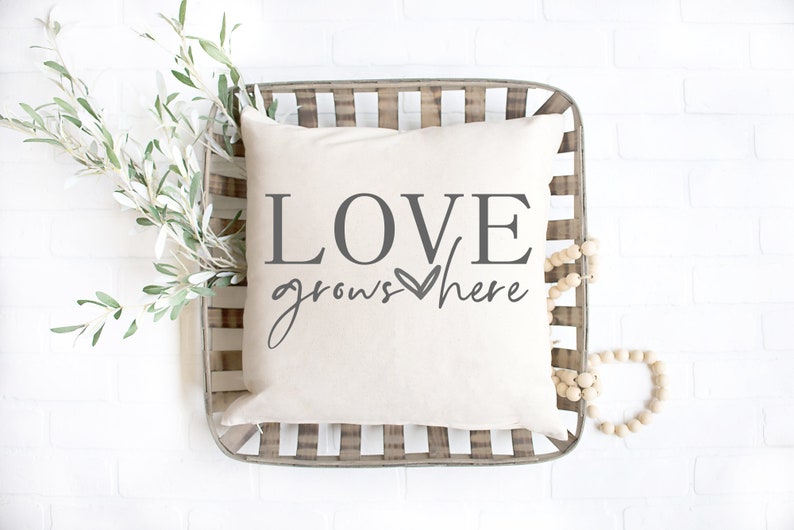 May include: A white throw pillow with the text "LOVE grows here" in gray lettering with a small heart symbol. The pillow is in a woven basket with a wooden bead garland.