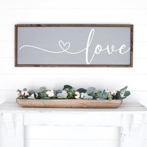 May include: A rectangular wooden sign with a grey background and the word "love" in white script, with a heart replacing the "o". Below, a wooden trough holds cotton and eucalyptus. The sign is 76 cm long.