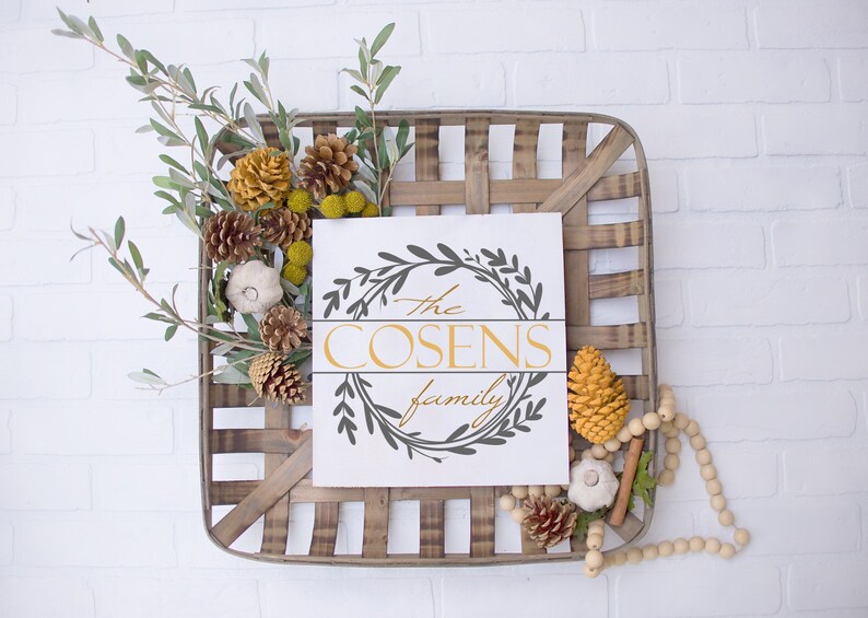 May include: A square, wooden basket with a white sign that reads "The Cosens family" in gold and gray. The basket is decorated with pinecones, faux pumpkins, and greenery. A string of wooden beads hangs from the basket.