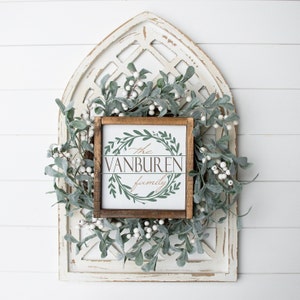 May include: A wooden framed sign with the text "The Vanburen Family" in a decorative font, surrounded by a green wreath design. The sign is set against a white distressed wooden background, with a green and white garland.