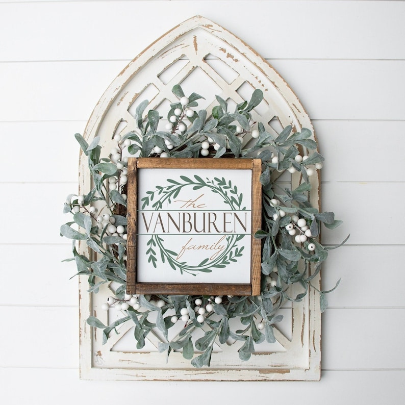 May include: A wooden framed sign with the text "The Vanburen Family" in a decorative font, surrounded by a green wreath design. The sign is set against a white distressed wooden background, with a green and white garland.