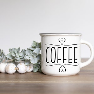 May include: White ceramic coffee mug with black rim and handle. The word "COFFEE" is written in black script with heart accents. The mug is on a wooden surface with decorative beads and greenery.