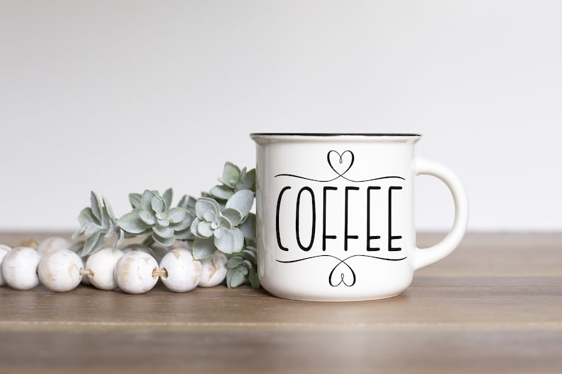 May include: White ceramic coffee mug with black rim and handle. The word "COFFEE" is written in black script with heart accents. The mug is on a wooden surface with decorative beads and greenery.
