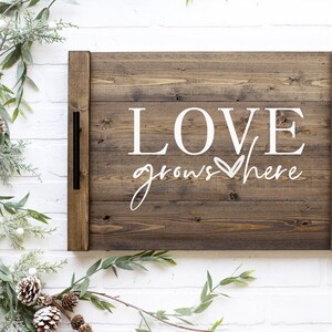 May include: A wooden tray with a brown stain and black metal handles. The tray has the words "LOVE grows here" written in white paint with a heart symbol.