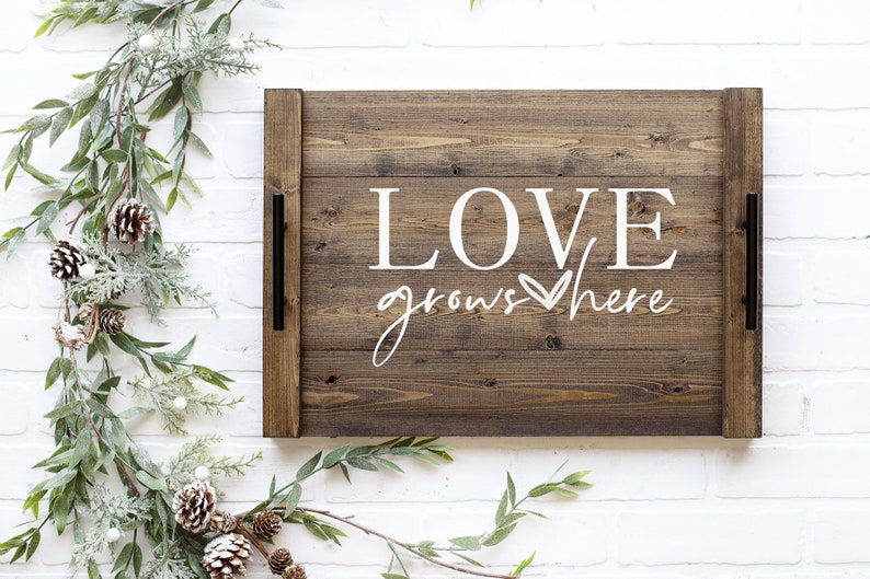 May include: A wooden tray with a brown stain and black metal handles. The tray has the words "LOVE grows here" written in white paint with a heart symbol.