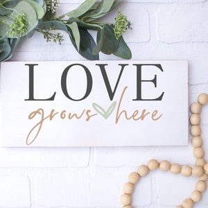 May include: White wooden sign with the text "LOVE grows here" in black and brown lettering with a green heart in the middle.
