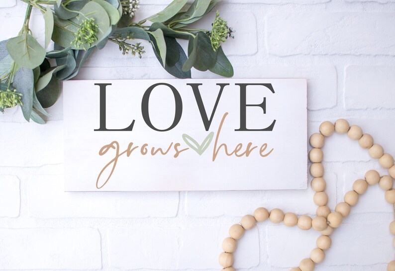 May include: White wooden sign with the text "LOVE grows here" in black and brown lettering with a green heart in the middle.