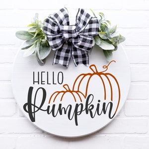 May include: A white round wooden sign with the text "Hello Pumpkin" in black script. Two orange pumpkins are drawn on the sign. A black and white checkered bow with green foliage is attached to the top of the sign.