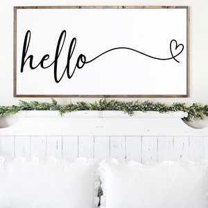 May include: A rectangular wooden framed sign with the word "hello" in cursive script, followed by a heart symbol. The sign is mounted above a white bed frame with white pillows and a green garland.