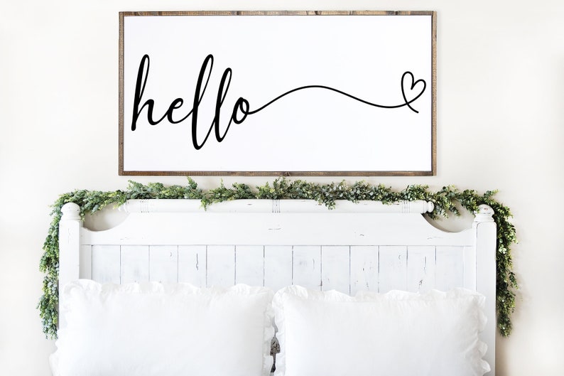 May include: A rectangular wooden framed sign with the word "hello" in cursive script, followed by a heart symbol. The sign is mounted above a white bed frame with white pillows and a green garland.