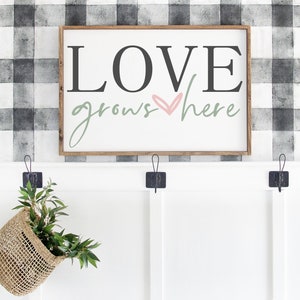 May include: A wooden sign with the text "LOVE grows here" in black and green lettering with a pink heart. The sign is hanging on a wall with a black and white checkered pattern.