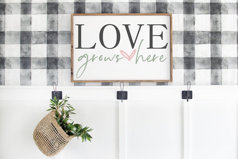 May include: A wooden sign with the text "LOVE grows here" in black and green lettering with a pink heart. The sign is hanging on a wall with a black and white checkered pattern.