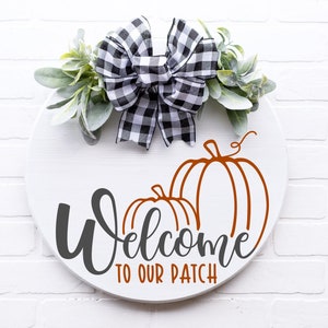 Op de afbeelding: Een rond wit houten bord met de tekst "Welcome to our patch" in zwarte schrijfletters. Twee oranje pompoenen staan op het bord en bovenop zit een zwart-wit geblokte strik met groen blad.