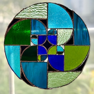 Puede incluir: Una vidriera con un diseño geométrico. La ventana presenta una forma circular con varias secciones de vidrio azul, verde y transparente. El vidrio está enmarcado con un borde oscuro. El diseño recuerda a una secuencia de Fibonacci.