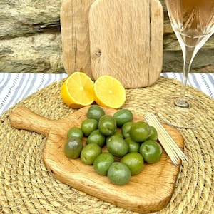May include: Two wooden chopping boards with handles, one is smaller than the other. The smaller board is filled with green olives and toothpicks. There are two lemon halves and a glass of rose wine in the background.