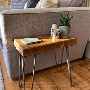 May include: A rustic wooden side table with hairpin legs. The table is made of reclaimed wood and has a natural finish. It is decorated with a small potted plant, a candle holder, and a coaster.