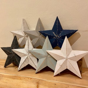 May include: A collection of six wooden star-shaped decorations in various shades of blue, grey, and white. The stars are arranged in a staggered pattern, with the largest star at the top and the smallest star at the bottom.