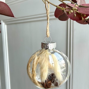 May include: A clear glass spherical ornament hanging from a jute twine loop. Inside the hollow sphere are beige dried bunny tail grasses and small brown pinecones. The ornament features a silver metal cap with a snowflake pattern.