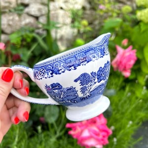 May include: A white ceramic creamer pitcher featuring a blue willow pattern with detailed illustrations of pagodas, trees, and bridges. The pitcher has a curved handle, a flared pouring spout, and a circular pedestal base with a glossy finish.