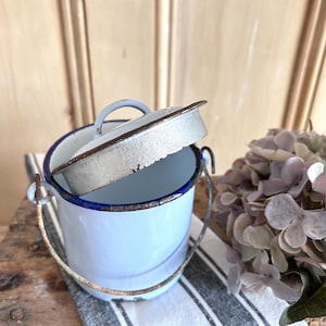 May include: A light blue enamel bucket with a dark blue rim and a matching lid. The bucket features a rusted metal swing handle and a small circular handle on the lid. The exterior finish shows signs of wear and aging with visible rust along the edges.