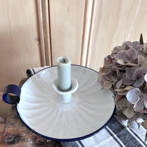 May include: A white enamel candle holder with a fluted design, a dark blue rim, and a matching blue loop handle. A single light gray taper candle sits in the center of the dish, which is positioned next to a cluster of dried hydrangea flowers.