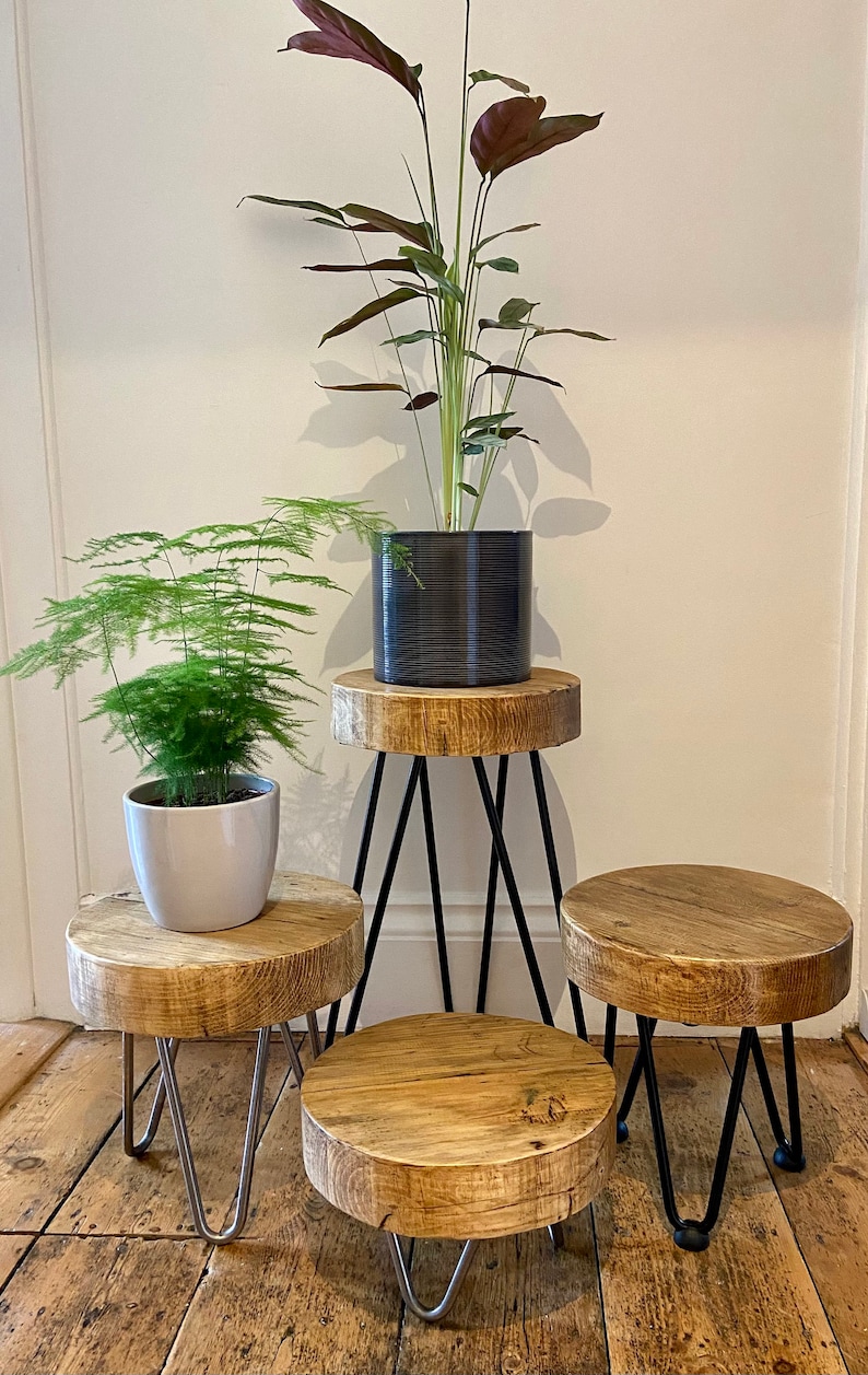 May include: Three wooden plant stands with black and silver hairpin legs. The stands are stacked on top of each other, with a potted plant on each stand. The top stand has a dark gray pot with a green plant with red leaves. The middle stand has a white pot with a green plant. The bottom stand is empty.