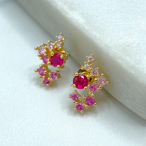 May include: A pair of gold-toned stud earrings. Each earring features a cluster of pink and fuchsia-colored gemstones, with a larger, round fuchsia stone at the center. The earrings are set against a white marble surface.