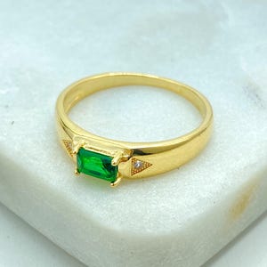 Puede incluir: Un anillo dorado con una piedra preciosa rectangular de color verde esmeralda. El anillo tiene una banda sencilla y pequeños detalles triangulares con pequeñas piedras transparentes a cada lado de la piedra principal. El anillo se muestra sobre una superficie blanca.