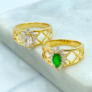May include: Two gold-colored rings with a geometric lattice design. One ring features a clear, oval-shaped stone surrounded by small, clear accent stones. The other ring has a green, oval-shaped stone with clear accent stones.