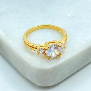 May include: A gold-colored ring with a large, clear, round center stone and smaller stones on either side. The ring has a detailed band with a textured design. The ring is set on a white surface.