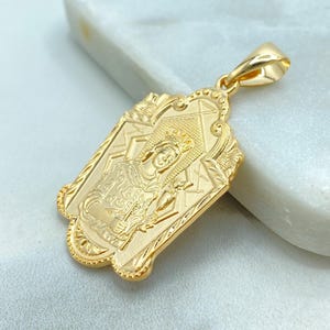 May include: A gold-coloured pendant with an ornate design featuring a religious figure. The pendant has a rectangular shape with a decorative border and a loop for a chain. The figure is detailed with intricate patterns and is set against a textured background.