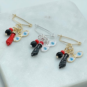 May include: Three gold safety pin brooches with dangling charms. Each brooch has a black hand charm, a red bead, and two blue evil eye charms.