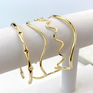 May include: Set of four gold-toned metal bangles with different designs. The bangles are stacked on a white display stand.