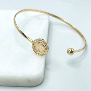 Puede incluir: Un brazalete de oro con un dije redondo que presenta una figura religiosa y una inscripción. El dije está unido al brazalete con una pequeña bola.