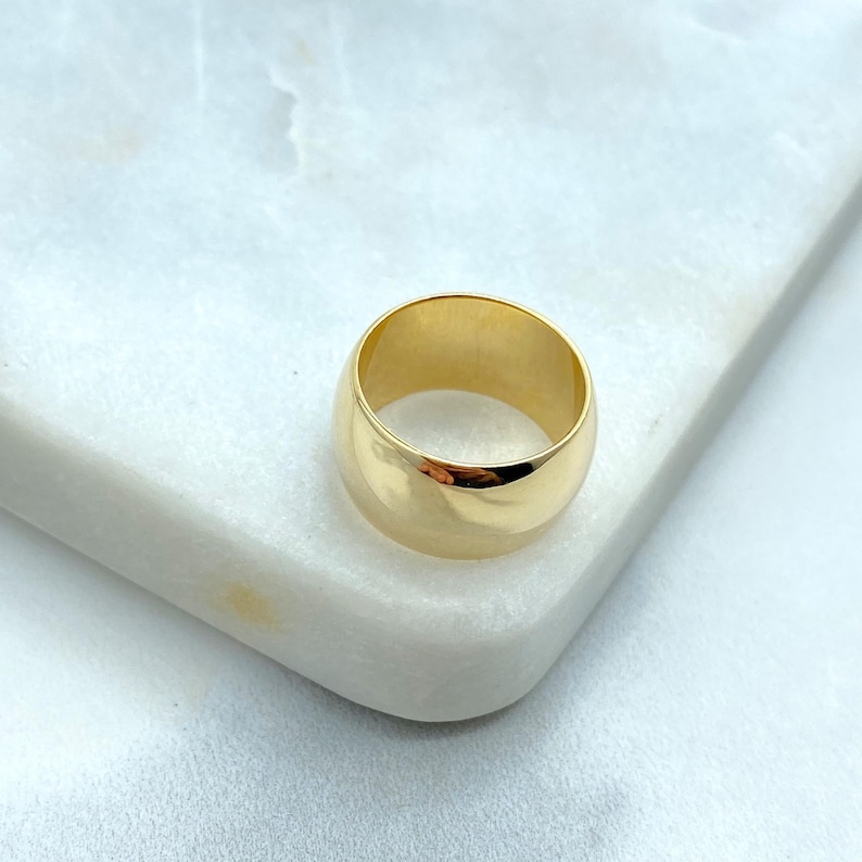 May include: A gold-colored ring with a wide band. The ring is smooth and shiny, reflecting light. The ring is set on a white marble surface. This ring is a piece of jewelry.