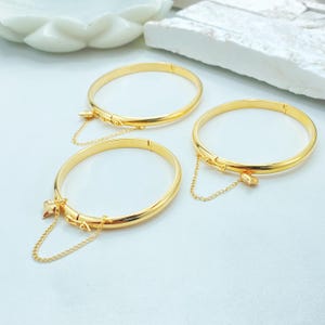 May include: Three gold-toned metal bangle bracelets, each featuring a smooth, polished finish and a hinged clasp mechanism. Attached to each bracelet is a delicate gold-toned chain with a small, heart-shaped charm dangling from the side.