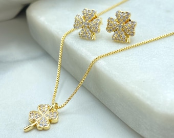 May include: A gold-colored jewelry set featuring a clover pendant necklace and matching stud earrings. The clover designs are embellished with small, clear gemstones. The set is displayed on a white surface.