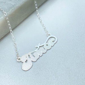 May include: A silver necklace featuring the word "Amor" in a cursive script. The pendant hangs from a fine chain. The necklace is presented against a light-toned backdrop.