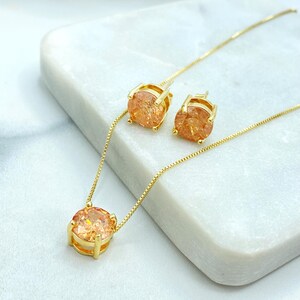 Könnte beinhalten: Ein goldfarbenes Schmuckset mit einer Halskette und passenden Ohrsteckern. Die Halskette hat eine feine Kette mit einem großen, runden, orangefarbenen Edelsteinanhänger. Die Ohrringe haben ebenfalls die gleichen orangefarbenen Edelsteine.
