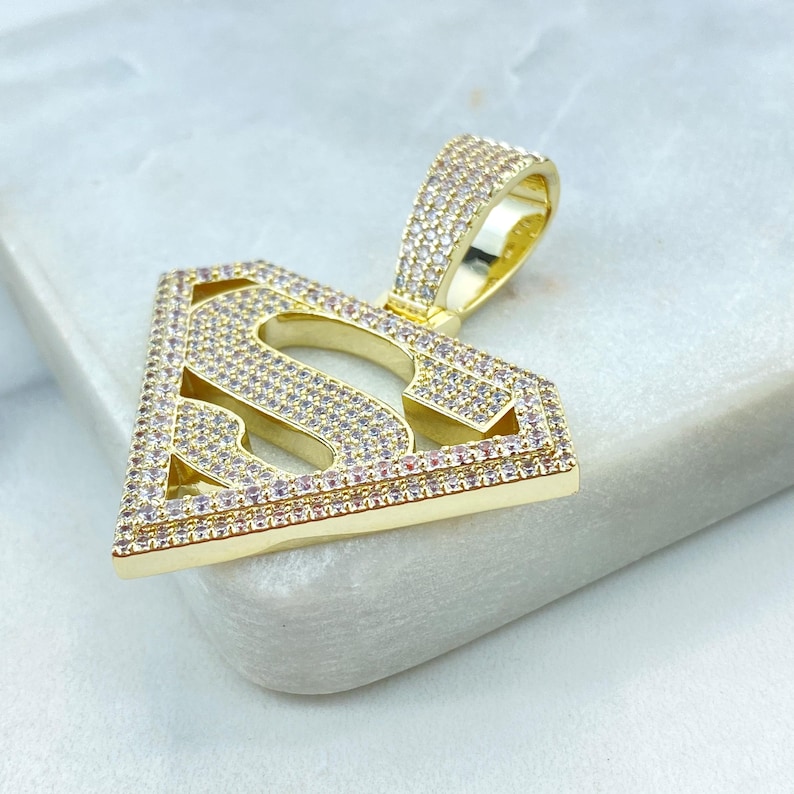 Puede incluir: Un colgante de tono dorado con un logotipo de Superman con un dise&ntilde;o de diamante. El colgante est&aacute; hecho de metal y tiene un acabado brillante.