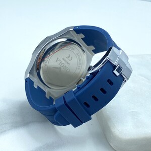 May include: A silver-toned wristwatch with a blue silicone band. The watch face is not visible. The back of the watch case has the words "ONOIA", "6098CH0", "CE", and "STAINLESS STEEL" printed on it.