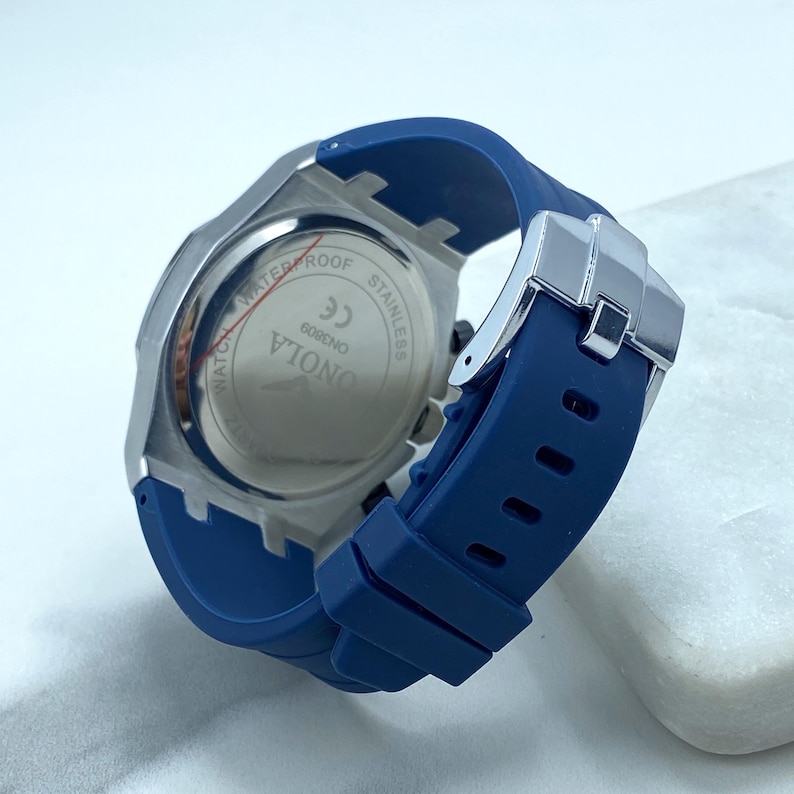 May include: A blue silicone wristband watch with a silver stainless steel case. The watch face is not visible in the image. The back of the watch case has the words "ONOIA 6080NO CE" and "WATERPROOF STAINLESS" printed on it.