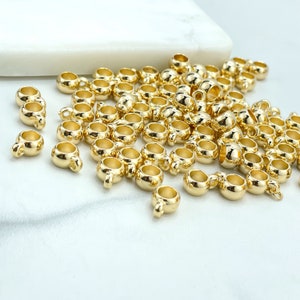 May include: A large group of gold-colored metal bead caps with a loop on the top. The bead caps are scattered on a white surface.