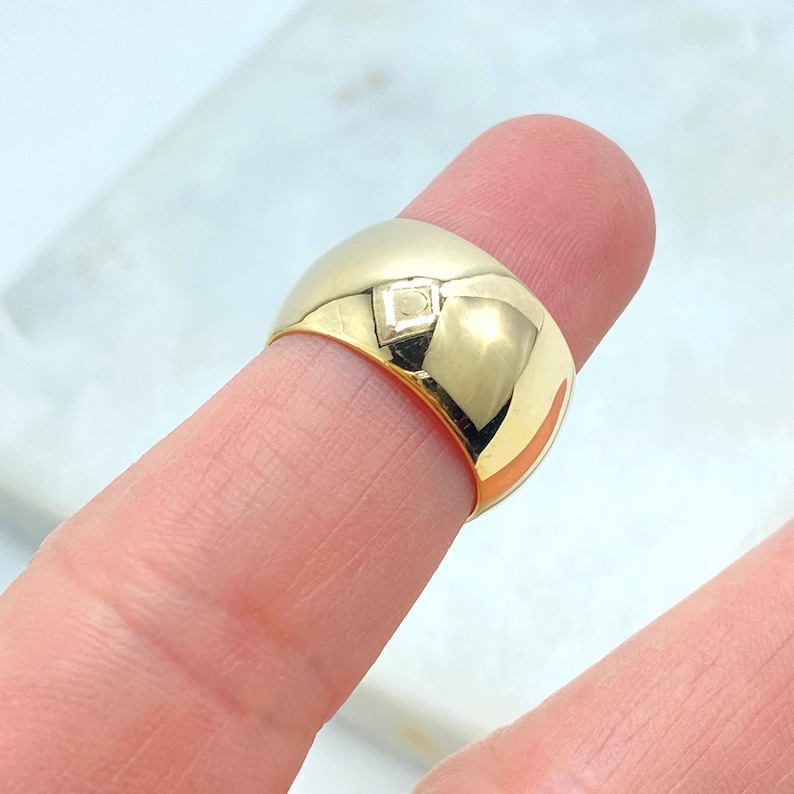 May include: A gold-colored ring with a rounded, domed shape. The ring has a small, square-shaped design element on the top. The ring is shown on a finger.