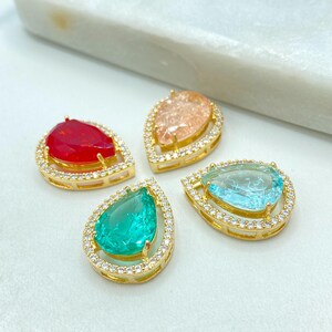 Puede incluir: Cuatro colgantes en forma de lágrima con engastes dorados y brillantes acentos transparentes. Los colgantes presentan piedras centrales de color rojo, melocotón, verde azulado y turquesa. Los colgantes se muestran sobre una superficie blanca.