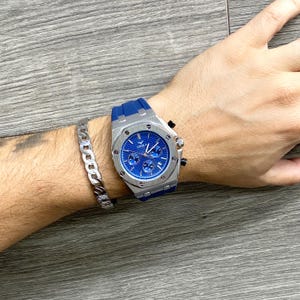 May include: A silver and blue chronograph wristwatch with a blue rubber band. The watch face has silver hands and markers, and the brand name "ONOLA" is printed on the face.